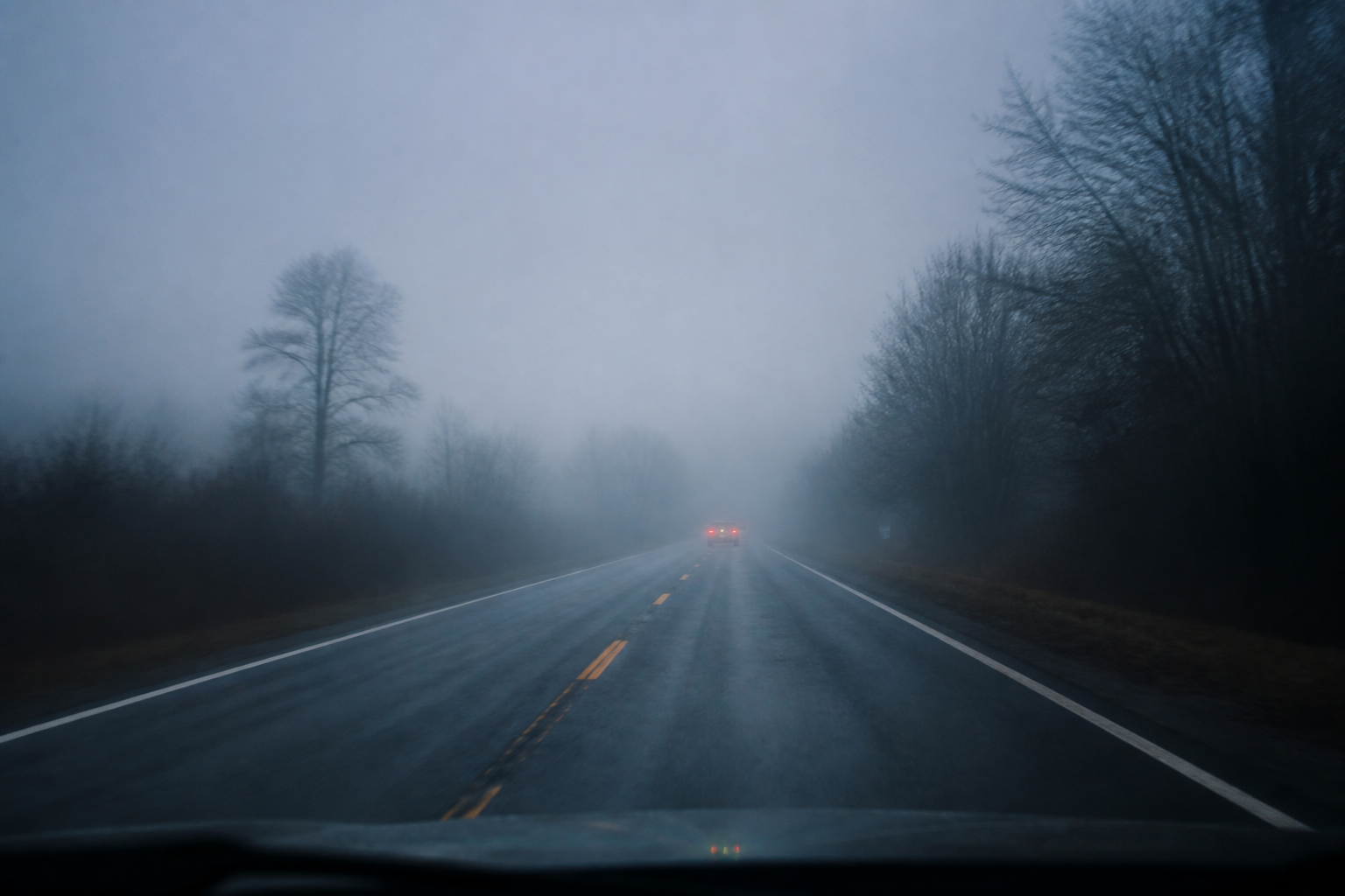 A foggy road at dawn with a car barely visible ahead